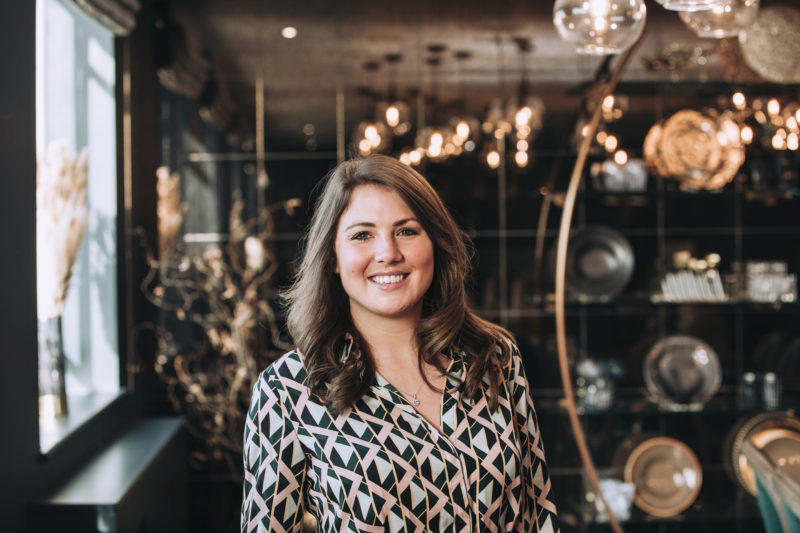 A woman with brown hair smiling in front of a blurred gold and bronze metal shelves with plates