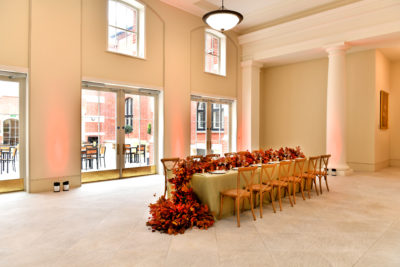 A long table decorated with autumnal leaves acting as a table runner with 6 chairs on each side in a large empty room at the Royal College of Music. There are double doors that lead to a patio with more table and chairs